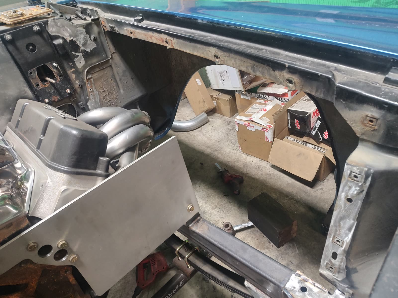 A classic car stripped to bare metal in the shop, a supercharger intake and hand-built stainless headers fitted, with a fabricated aluminium panel bolted into the engine bay.