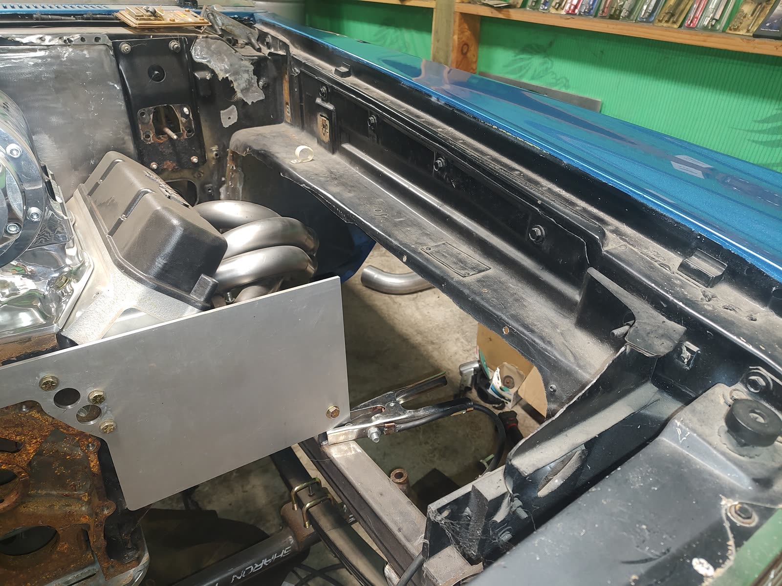 Engine bay fabrication in progress on a blue classic car, the welding ground clamp still clipped to the panel.